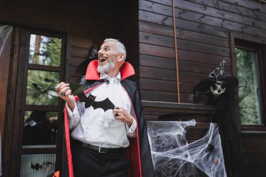 Vampir cadılar bayramı kostümü giymiş olgun bir adam dekore edilmiş evin verandasında gülüyor.