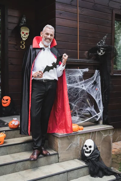 happy mature man in vampire costume holding paper cut bat on porch with halloween decoration