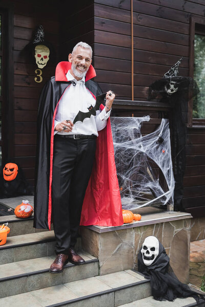 счастливый зрелый мужчина в костюме вампира, держащий бумагу порезанную летучей мышью на крыльце с украшением на Хэллоуин
