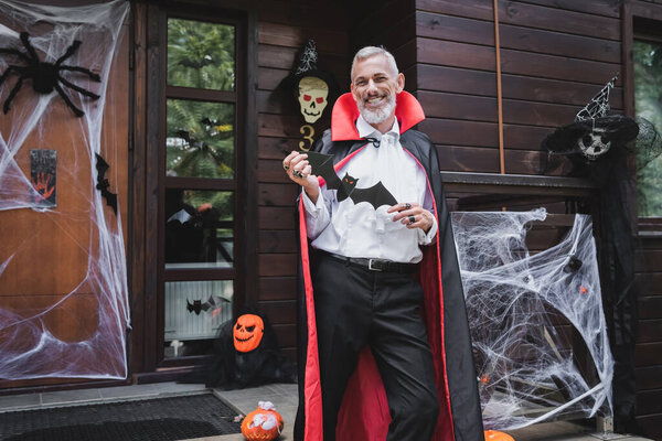 счастливый зрелый мужчина в костюме вампира Хэллоуина, стоящий на украшенном крыльце с отрезанной папочкой битой