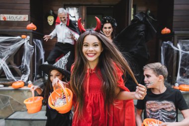 happy girl in devil halloween costume holding bucket of sweets while running out neighbor cottage with friends