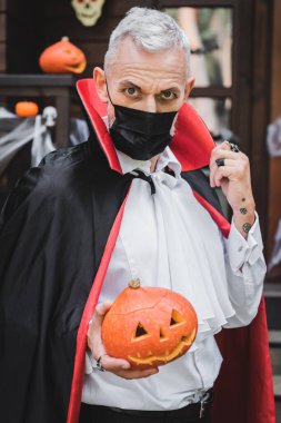 middle aged man in black medical mask and vampire halloween costume holding carved pumpkin while looking at camera  clipart