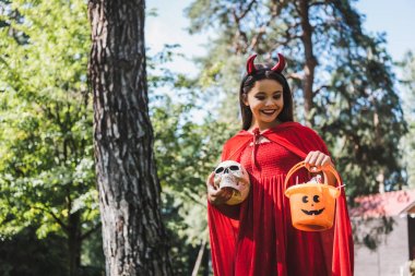 smiling child in devil halloween costume holding skull and bucket with candies in forest clipart