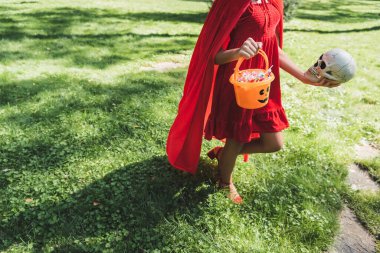 Kırmızılı cadılar bayramı kostümlü çocuk kafatası ve şeker dolu kovayla çimlerde yürüyor.