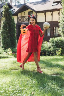 cheerful girl in devil halloween costume walking with skull and bucket of sweets near blurred cottage clipart