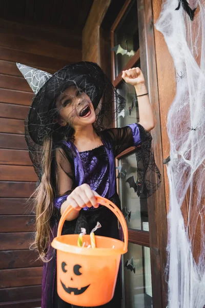 excited girl in witch hat knocking at door while holding bucket with sweets