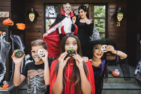 excited kids in scary costumes having fun with halloween cookies while mother waving hand on blurred background