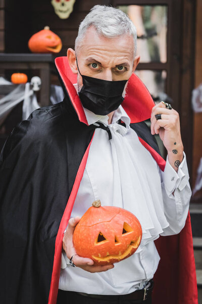 middle aged man in black medical mask and vampire halloween costume holding carved pumpkin while looking at camera 