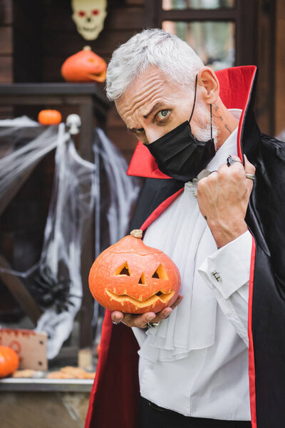 mature man in vampire halloween costume and black medical mask looking at camera while holding carved pumpkin