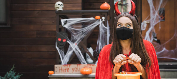 excited girl in devil costume and black medical mask holding bucket with candies near blurred decorated house, banner