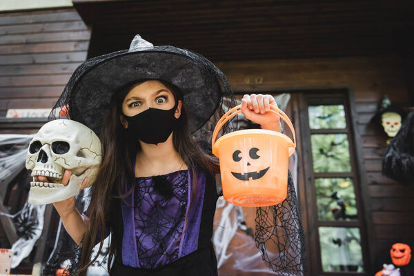 child in witch halloween costume and black medical mask holding skull and bucket while looking at camera