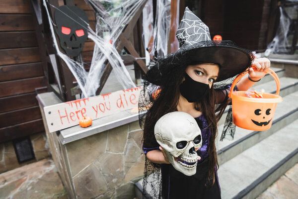 girl in witch halloween costume holding skull and bucket of sweets near card with here you die lettering