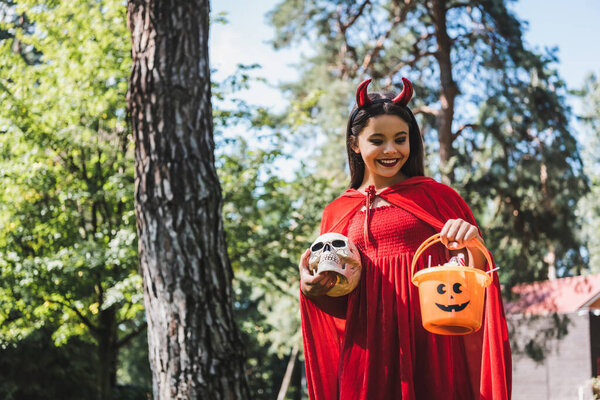 smiling child in devil halloween costume holding skull and bucket with candies in forest