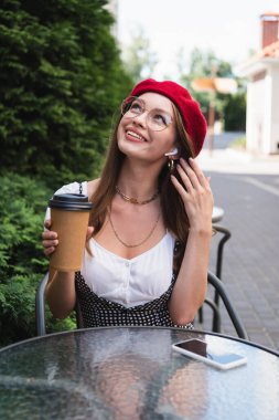 Kırmızı bereli mutlu genç bir kadın, kulaklıklar ve gözlüklerle akıllı telefonun yanında boş ekranlı kağıt bardak tutuyor. 