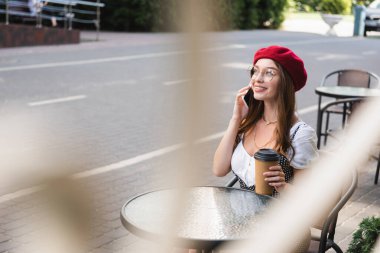 Kırmızı bereli, gözlüklü, elinde kağıt bardakla gülümseyen ve kafenin terasında akıllı telefondan konuşan bir kadın.