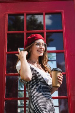 Bereli mutlu bir kadın ve kırmızı telefon kulübesinin yanında kağıt bardak tutan gözlüklü bir kadın.