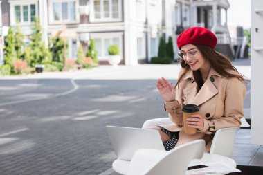 Kırmızı bereli ve bej renkli trençkotlu mutlu genç kadın kafenin terasında el sallıyor. 