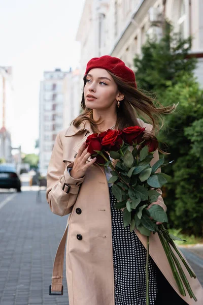 pretty woman in trench coat and red beret holding bouquet of roses on urban street of europe