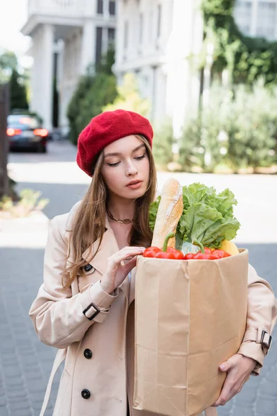 young woman in beige trench coat and red beret looking at paper bag with groceries on urban street