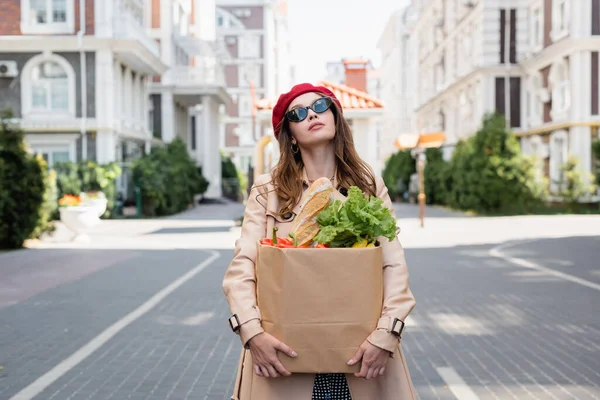ベージュのトレンチコートを着た夢のような女性サングラスと赤いベレー帽ヨーロッパの都市部の通りに食料品と紙の袋を保持 — ストック写真