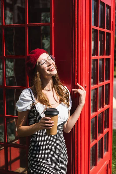 Mujer Alegre Boina Anteojos Sosteniendo Taza Papel Cerca Cabina Telefónica — Foto de Stock