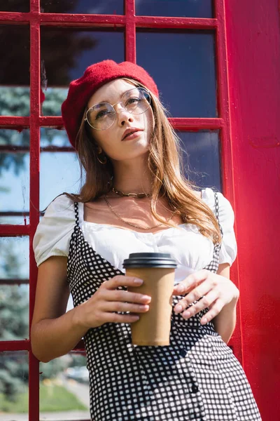 戴着贝雷帽 戴着眼镜 拿着纸杯 站在红色电话亭旁的漂亮女子 — 图库照片
