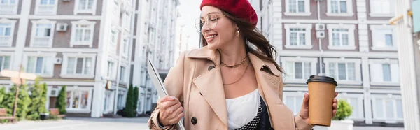 赤いベレー帽とベージュのトレンチコートを着た楽しい女性ノートパソコンとストリートの紙コップ バナー — ストック写真