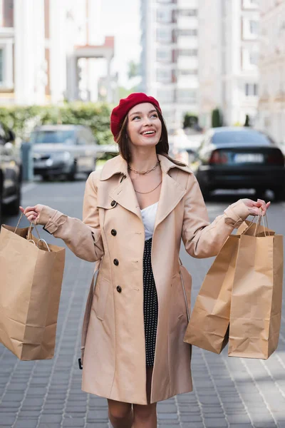 cheerful woman in red beret and beige trench coat holding shopping bags on urban street