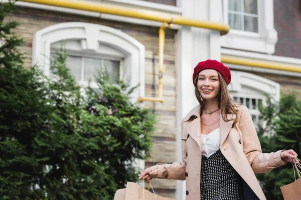 young happy woman in red beret and beige trench coat holding shopping bags outside