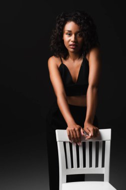 Baştan çıkarıcı Afro-Amerikalı kadın saten bluzlu. Siyah sandalyenin yanında poz veriyor. 