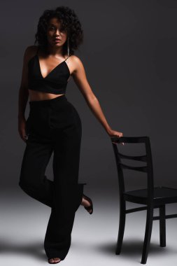 Esmer, Afro-Amerikalı, seksi elbiseli, koyu gri sandalyenin yanında poz veren bir kadın.