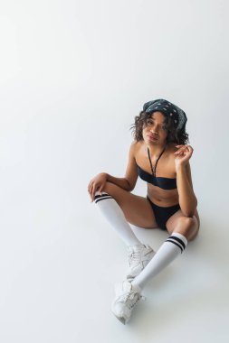 Yüksek açılı, şık Afro-Amerikan kadın mayo, diz boyu çoraplar ve gri ayakkabılarla oturuyor.