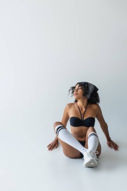 Uzun boylu, şık Afro-Amerikan kadın mayo, diz boyu çoraplar ve gri ayakkabılarla oturuyor.