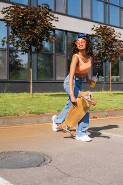Afro-Amerikan kadın, güneş gözlüklü, elinde portakal suyu ve dışarıda uzun pano olan plastik bir bardak tutuyor. 