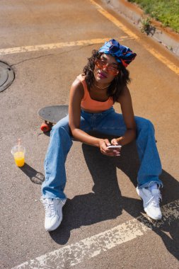 Afrika kökenli, güneş gözlüklü, uzun panoda oturan ve asfalttaki plastik bardağın yanında akıllı telefon kullanan Amerikalı kadının yüksek açılı görüntüsü. 