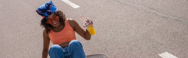 Güneş gözlüklü mutlu Afro-Amerikalı kadın elinde portakal suyuyla plastik bardakla dışarıdaki panoda oturuyor.