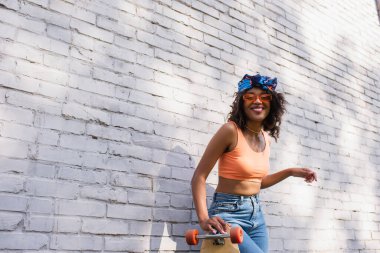 Güneş gözlüklü ve başörtülü neşeli Afro-Amerikan kadın tuğla duvarın yanında uzun pano tutuyor.