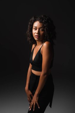 Şık Afro-Amerikan kadın saten bluz ve pantolon giymiş, siyah üzerinde poz veriyor. 