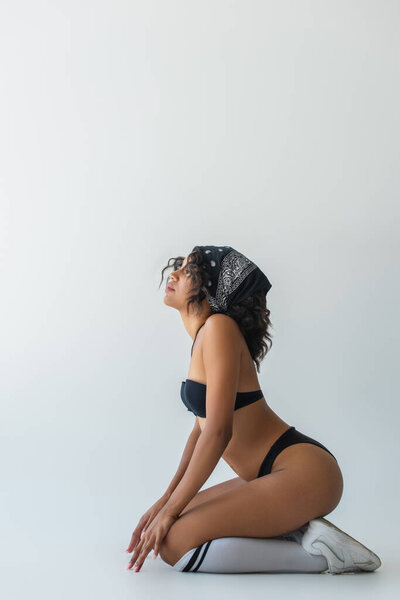 Вид сбоку на стильную африканскую женщину в купальнике, с высоко поднятыми коленями и позирующей на сером фоне