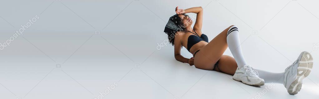 full length of stylish african american woman in swimsuit, knee high socks and sneakers lying on grey, banner