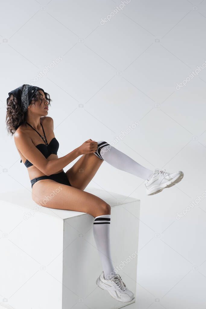 full length of sexy african american woman in swimsuit and sneakers adjusting knee high socks while sitting on cube isolated on grey