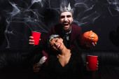 laughing man in vampire halloween costume with plastic cup and carved pumpkin near african american woman in wolf mask on black  
