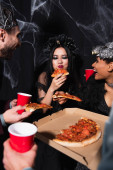potěšený asijské žena v upír halloween kostým jíst pizza poblíž multietnické přátelé na černý
