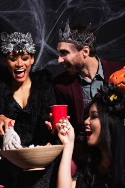 excited asian woman taking popcorn with toy hand near multiethnic friends in halloween costumes on black clipart