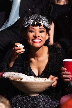 excited african american woman in wolf mask eating popcorn during halloween party on black clipart