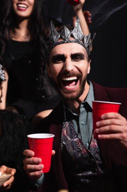 Vampir kralın tacındaki adam siyah arka planda bulanık bir kadının yanında plastik bardak tutuyor.