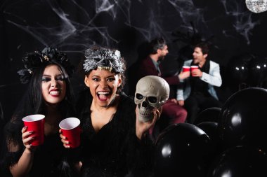 interracial women in spooky halloween costumes grinning and growling near blurred friends talking on black   clipart