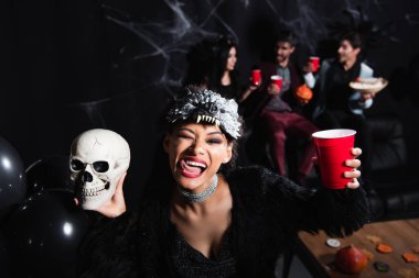 african american woman in wolf halloween mask sticking out tongue while holding spooky skull and plastic cup on black  clipart