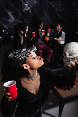 african american woman in werewolf halloween mask pouting lips while looking at creepy skull on black clipart