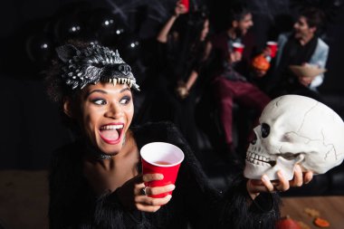 amazed african american woman in wolf mask looking at scary skull near blurred friends on black background clipart